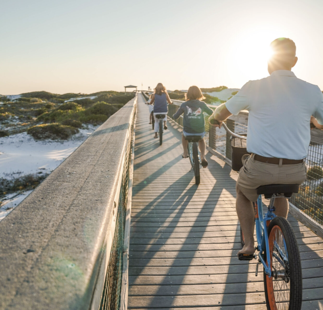A smiling family riding colorful beach cruiser bikes along a wooden boardwalk with ocean views, showcasing 30A bike rentals for a fun and scenic way to explore Florida’s Emerald Coast.